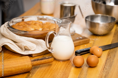 Tasty apple pie on a oaken wood table with a ingredients. Woman in a gray pajama cooking a delicious apple pie on a kitchen in a loft style. Milk, eggs, sugar and a baked pie on a table. Home kitchen.