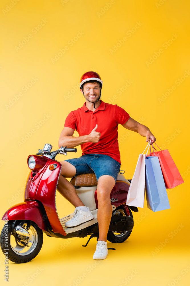 happy delivery man in helmet sitting on scooter and holding shopping bags while showing thumb up on yellow