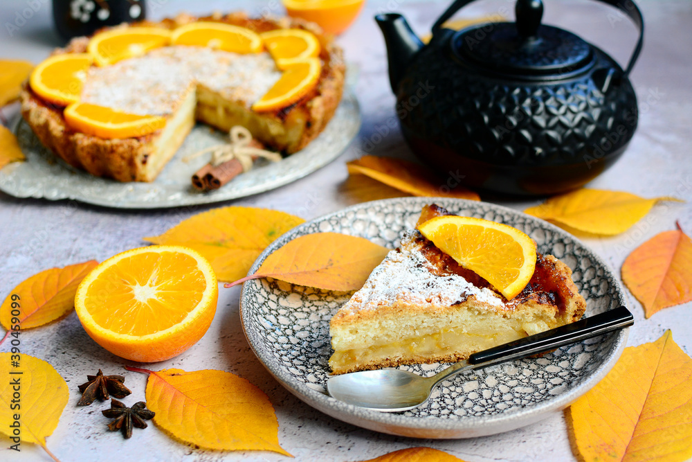A piece of lemon pie on a plate, oranges, yellow leaves, a teapot on a light gray table. Autumn still life with dlmashny pastries. A piece of lemon pie on a plate, oranges, yellow leaves, a teapot on a light gray table. Autumn still life with dlmashny pastries.