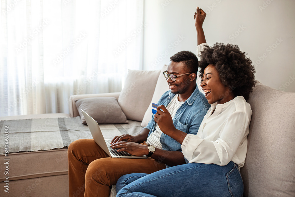 Overjoyed african American couple sit relax on cozy couch look at laptop screen triumph winning lottery online, happy biracial husband and wife feel excited euphoric with good news on computer