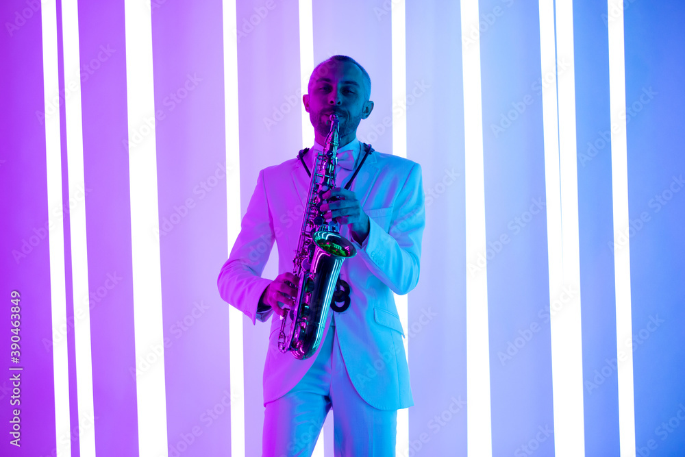 Fototapeta premium Happy saxophonist plays music on sax in elegant white suit. Man stand in the studio against the backdrop of neon lamps with blue backlight.
