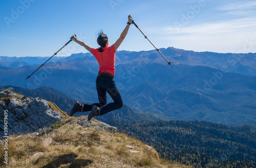 happy woman hiker goes hiking, jumps on top of mountain and looks at autumn nature mountains landscape in sunny weather. Outdoor activities. Copy space, back view, hands up