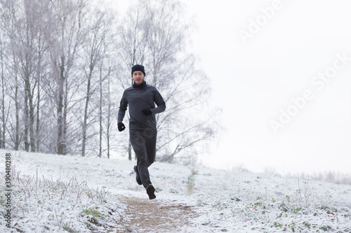 Young, adult alone man in sport tracksuit and hat running on park trail in winter day after first snow. Front view. 