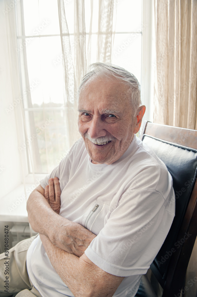 old man is sitting in a chair. Smiling old man sitting in a chair by the window, portrait, close-up old man is sitting in a chair. Smiling old man sitting in a chair by the window, portrait, close-up