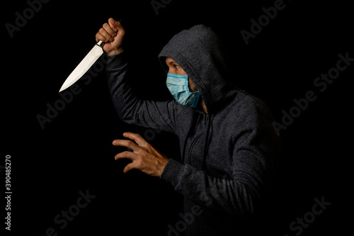 Aggressive young man in a medical mask in a hood with a knife on a black background.