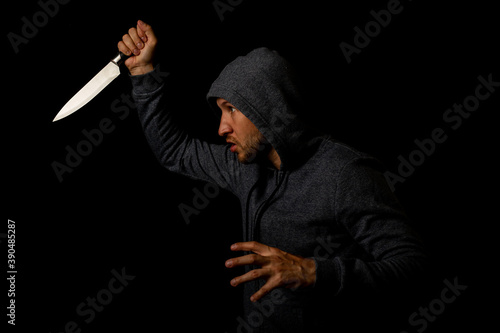 Aggressive young man in a hood brandishes a knife against a black background.