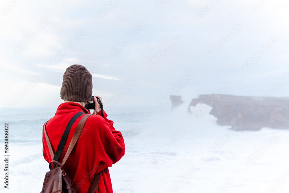 An unrecognizable photographer from the back, wearing a red sweatshirt and wool hat, takes pictures in a passage of sea cliffs. Bring a backpack. hobbies and free time concept.