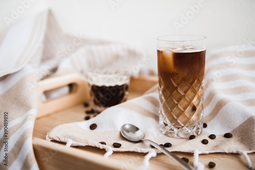 Black ice coffee with milk in the vintage glass on the ligh background, wooden tray and beige striped tablecloth, coffee beans
