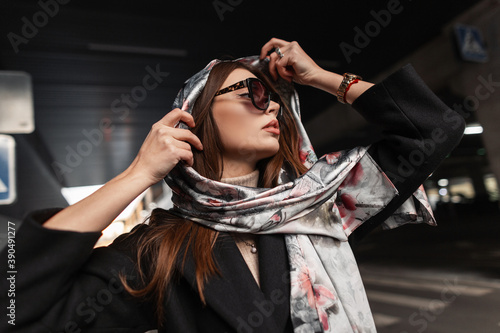 Elegant young pretty beautiful woman in stylish dark sunglasses in a black coat puts a luxurious silk scarf on head outdoors. Beautiful girl fashion model relaxes on the road in the city. Sexy lady. photo