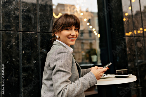 Happy positive woman dressed grey jacket posing at camera with smartphone on outside cafe with lights