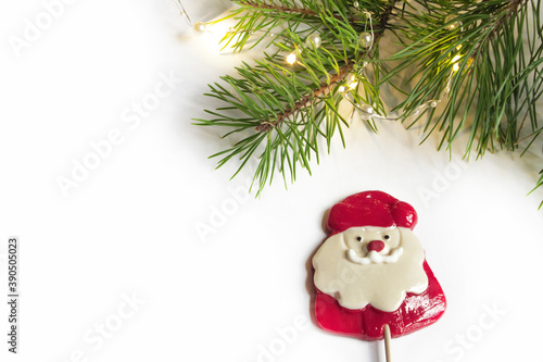 Christmas composition of a natural pine branch with a Christmas composition of a natural pine branch with a garland and a Lollipop in the form of Santa Claus, light background. Corner, frame for banne