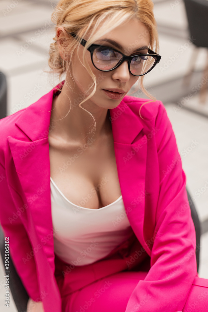 Pretty sexy young woman fashion model with big breasts in stylish pink suit  in fashionable glasses is sitting in office. Beautiful girl blonde in  trendy glamorous outfit resting indoors. Beauty lady. Stock