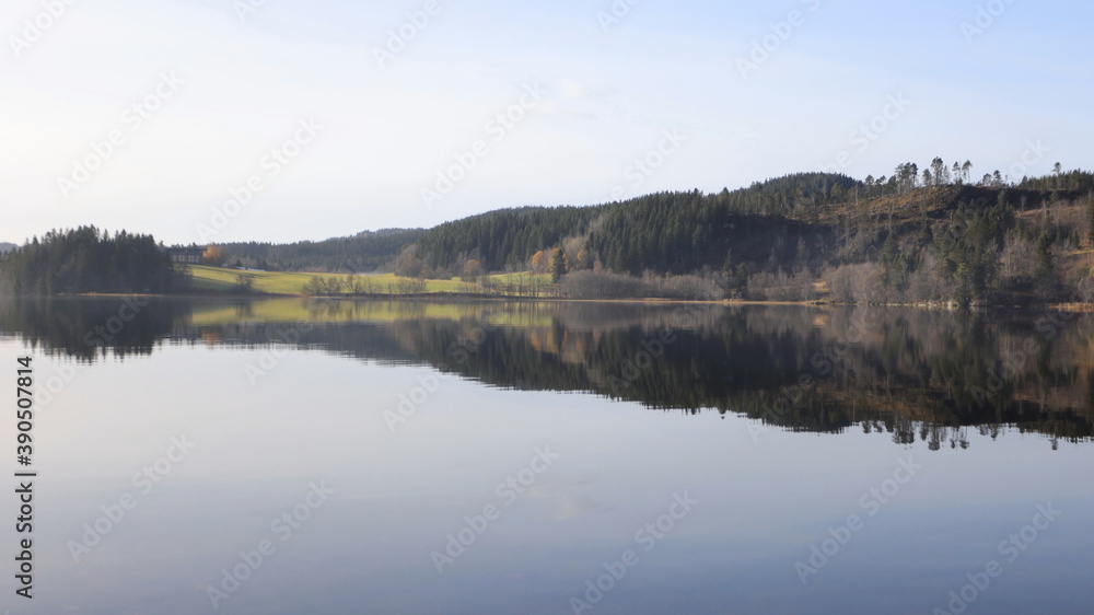 Fototapeta premium Grassland, woods and farmland in dawn, with mist in a sunny morning. The early autumn reflection along lakeshore reflected in the water.