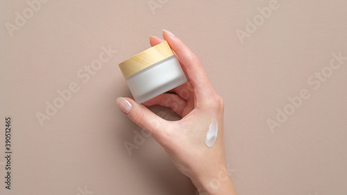 Female hand holding jar of cosmetic cream on pastel beige background. Woman applying moisturizer cream photo