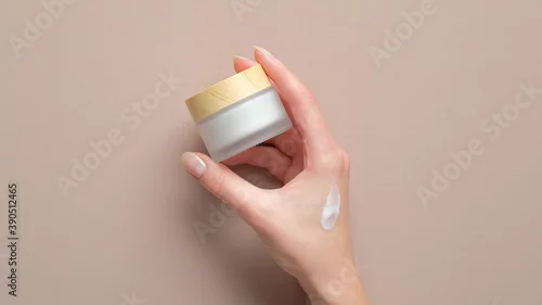 Obraz Female hand holding jar of cosmetic cream on pastel beige background. Woman applying moisturizer cream