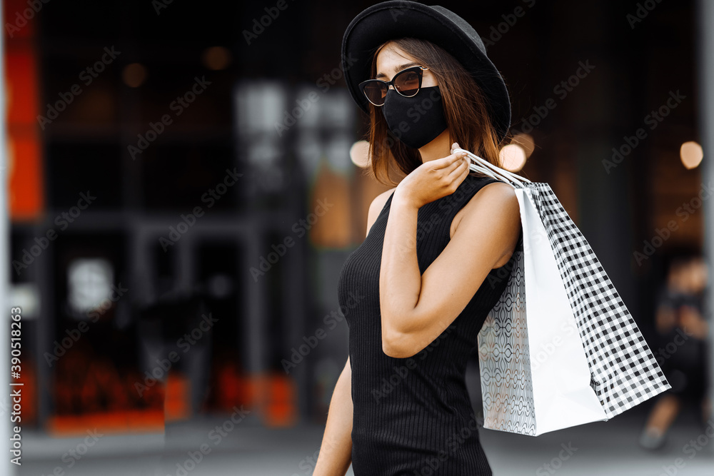 stylish Shopaholic girl in a medical protective mask on her face, in a black dress and hat, shopping in a shopping center, with shopping bags. Black Friday, shopping
