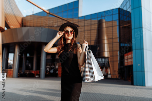 Happy attractive elegant girl, in a black dress and hat, wearing dark glasses, holding shopping bags and enjoying shopping. Black Friday, shopping
