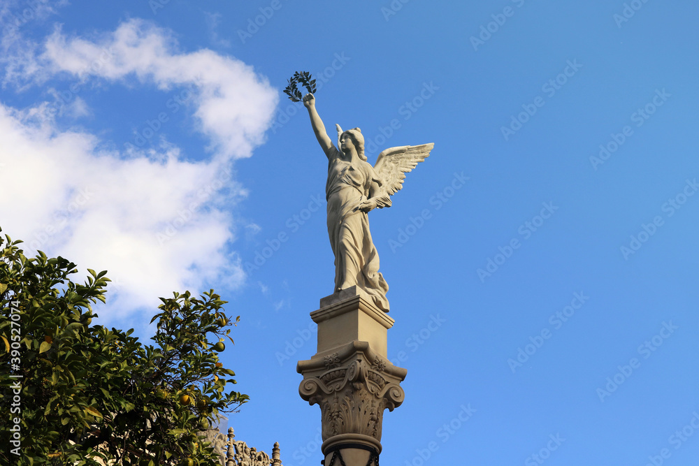 Statue of a female angel with wings and a laurel wreath in the hand, with a blue sky in the background, a tree and clouds 