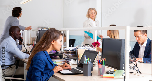 Young serious businesswoman concentrated on work with laptop in coworking space with international team