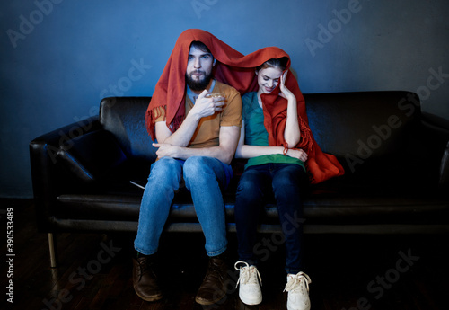a woman and a man under a red blanket on the couch watching tv in the evening