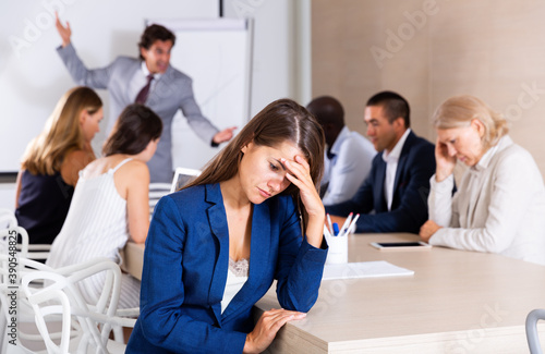 Young woman sitting in meeting room looking upset because of angry boss scolding her coworkers