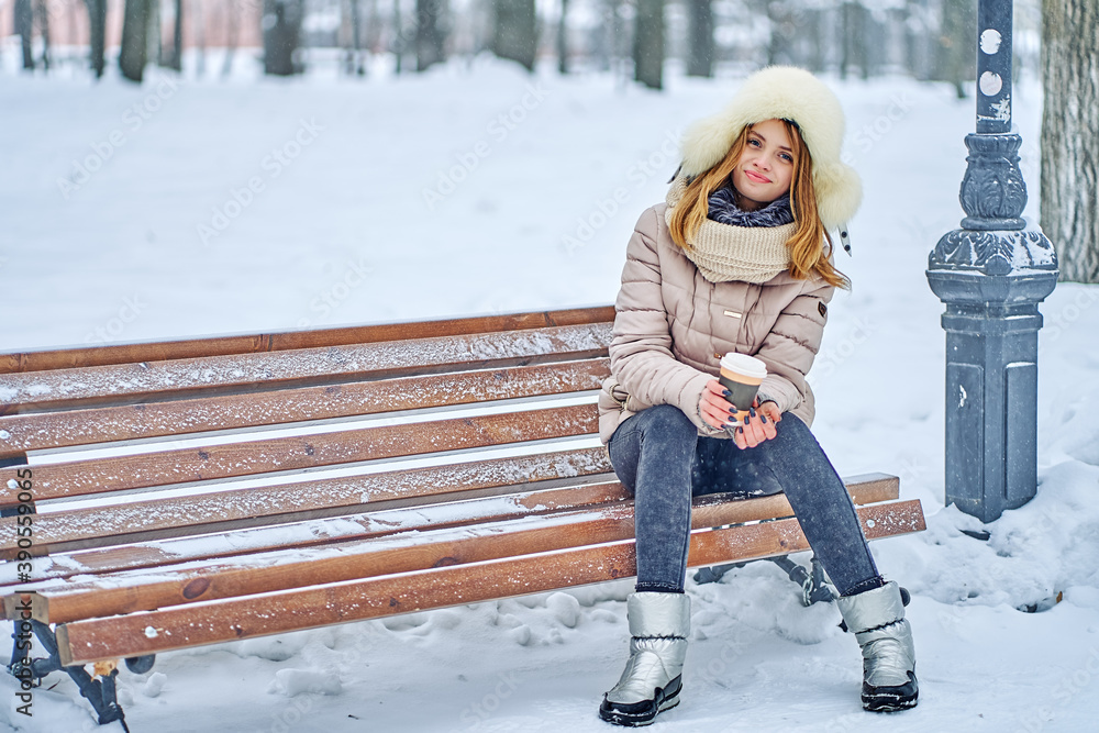Attractive woman sits on a bench in a winter park. She is dressed warmly. In hand is a paper cup with hot coffee. copy space - obrazy, fototapety, plakaty 