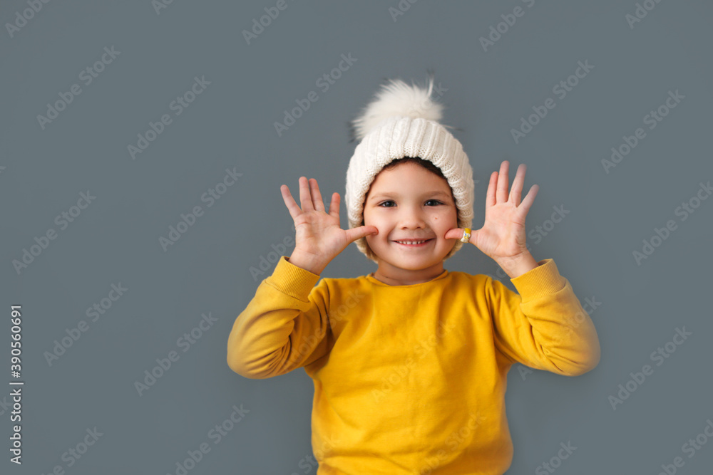Funny crazy caucasian child girl in white knitted hat and yellow sweater on a gray background, the concept of childish emotions and antics. Funny crazy caucasian child girl in white knitted hat and yellow sweater on a gray background, the concept of childish emotions and antics.
