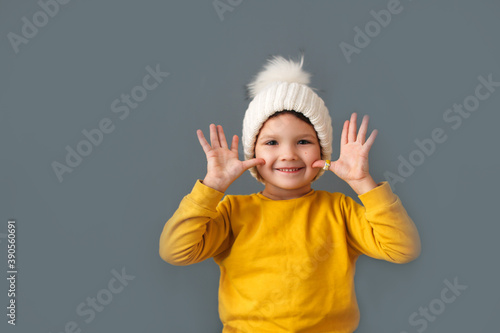 Funny crazy caucasian child girl in white knitted hat and yellow sweater on a gray background, the concept of childish emotions and antics.