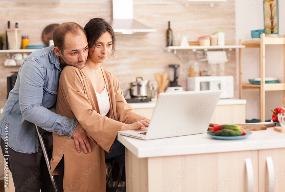 Wife reading on laptop in kitchen while husband is hugging her. Happy loving cheerful romantic in love couple at home using modern wifi wireless internet technology Wife reading on laptop in kitchen while husband is hugging her. Happy loving cheerful romantic in love couple at home using modern wifi wireless internet technology