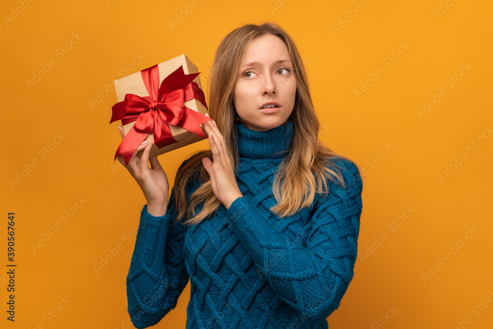 Portrait of a puzzled young woman holding gift decorated with ribbon. Studio shot, yellow background Portrait of a puzzled young woman holding gift decorated with ribbon. Studio shot, yellow background