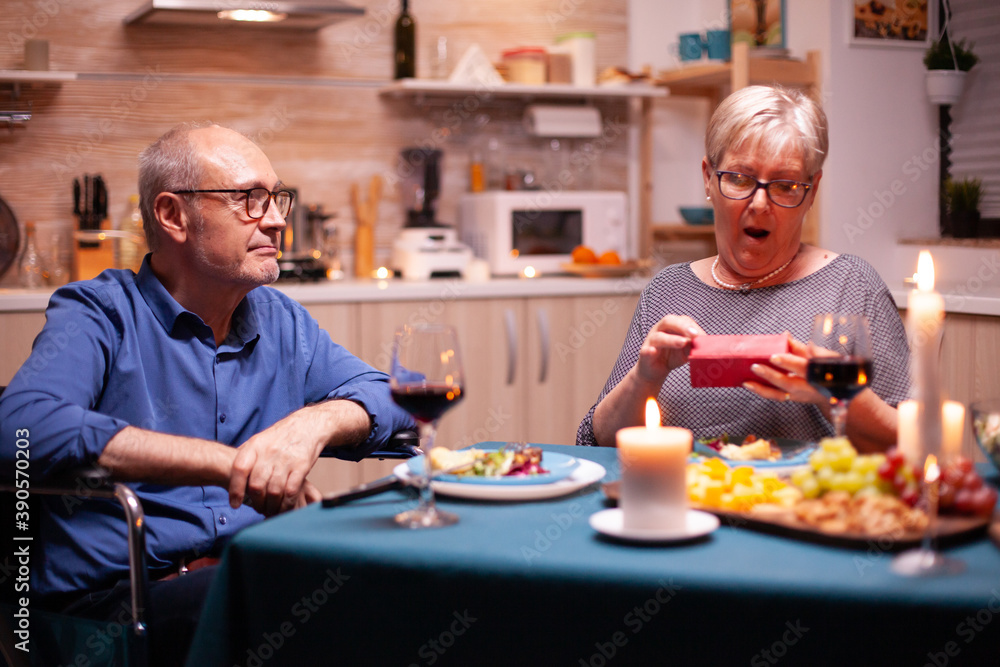 Old woman looking surprised holding gift from husband in wheelchair during festive dinner. Happy cheerful elderly couple dining together at home, enjoying the meal, celebrating their marriage ,