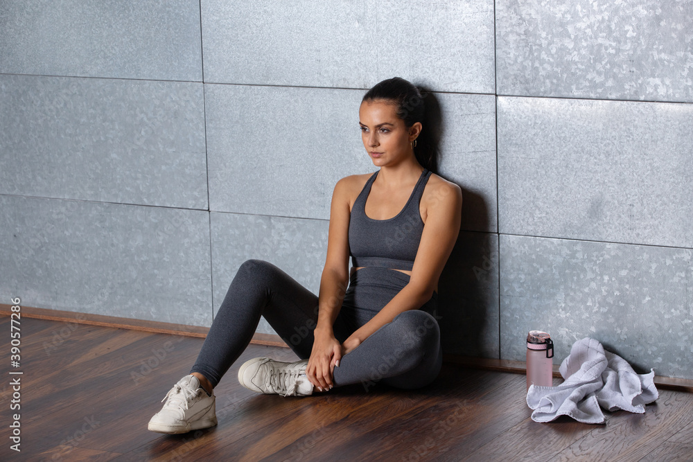Fototapeta premium Female fitness trainer sitting on the floor on a metal wall with her bottle and a towel in fitness outfit with athletic shape