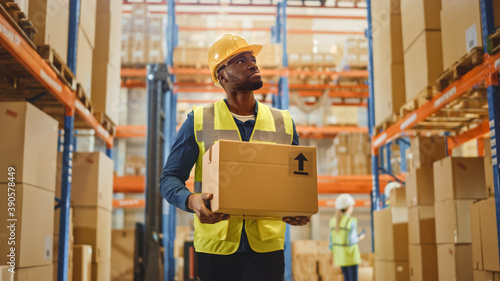 Handsome Male Worker Wearing Hard Hat Holding Cardboard Box Walking Through Retail Warehouse full of Shelves with Goods. Working in Logistics and Distribution Center. Front Shot.