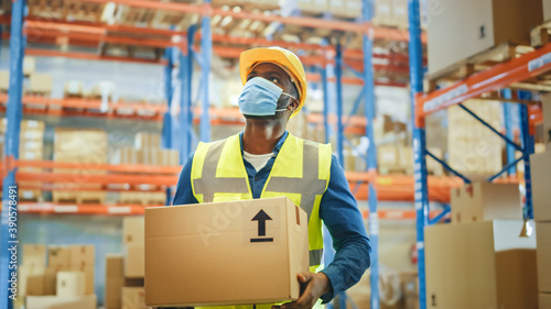 Portrait of Handsome Male Worker Wearing Medical Face Mask and Hard Hat Carries Cardboard Box Walks Through Retail Warehouse full of Shelves with Goods. Safety First Protective Workplace.