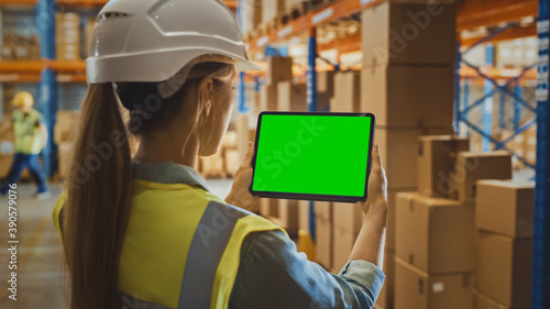 Professional Female Worker Wearing Hard Hat Uses Digital Tablet Computer with Green Chroma Key Screen in Landscape Mode in the Retail Warehouse full of Shelves with Goods. Over the Shoulder view 