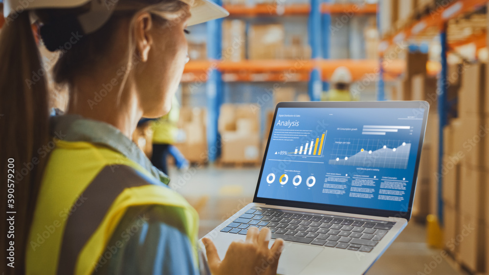 © Gorodenkoff - Professional Female Worker Wearing Hard Hat Holds Laptop Computer with Screen Showing Analysis Software in the Retail Warehouse full of Shelves with Goods. Over the Shoulder Side View