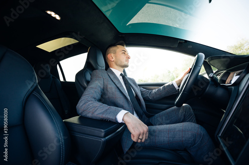 Portrait of male businessman sitting at the wheel of a car. Young cheerful man sitting in modern luxurious car staying in city center