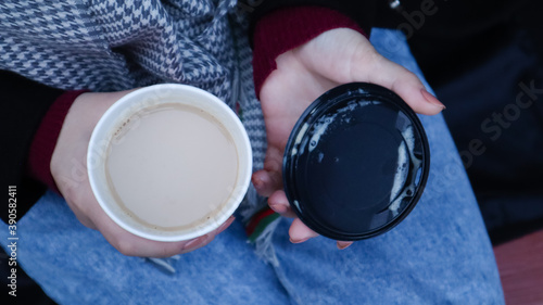 Female hands are holding morning takeaway coffee and closing a paper cup with a lid while sitting on a park bench, close-up of hands.