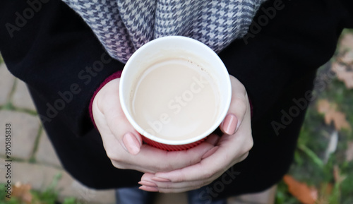 A young girl in a coat holds morning coffee with her while walking in the park. Hand holding paper cup of coffee in green park. Takeaway cappuccino. Close-up, top view.
