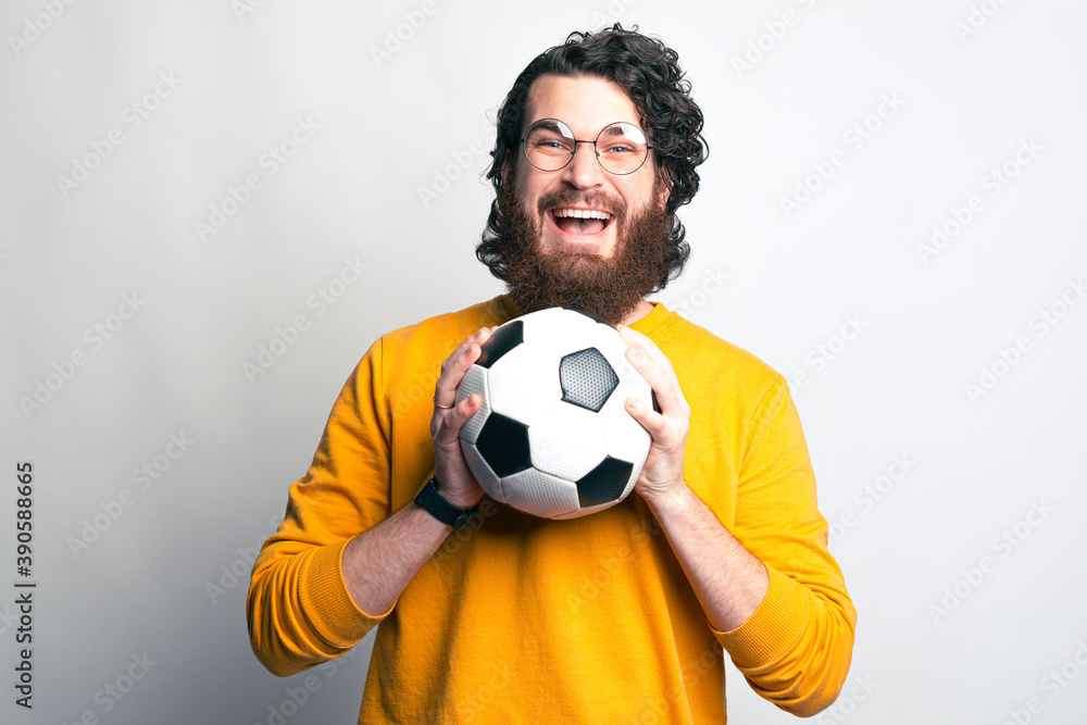 A bearded man with glasses is looking excited at the camera holding a soccer ball with both hands .
