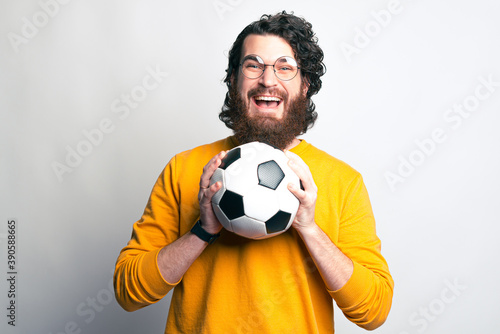 A bearded man with glasses is looking excited at the camera holding a soccer ball with both hands . © Vulp