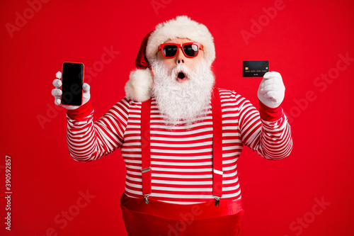 Portrait of his he handsome amazed stunned bearded fat overweight Santa holding in hands gadget bank card order app shopping service sale isolated bright vivid shine vibrant red color background © deagreez
