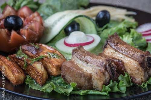 Bacon grill and fried potatoes on lettuce leaves with tomato and cucumber in a black plate