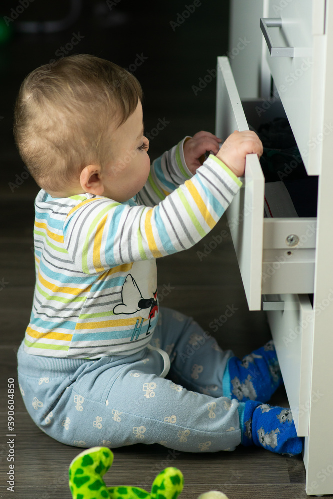 Little caucasian baby boy opening a white drawer with underwear when nobody is watching. Naughty toddler exploring dangerous furniture. Baby safe at home concept.