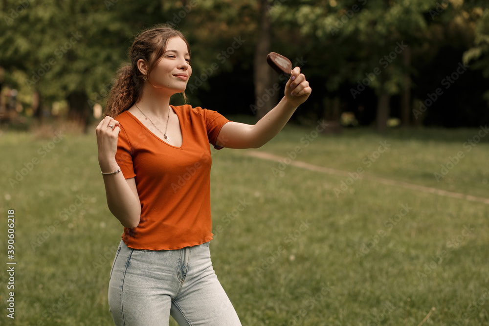 Horizontal portrait of young smiling girl college student holding ice lolly cream in hand. Woman dressed in orange t-shirt in sunny weather standing in the park. Nature blur background, copy space. Horizontal portrait of young smiling girl college student holding ice lolly cream in hand. Woman dressed in orange t-shirt in sunny weather standing in the park. Nature blur background, copy space.