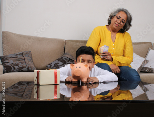 Indian/asian Grandma teaching her grand son importance of savings, putting coins into pink piggy bank.