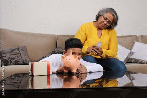 Indian/asian Grandma teaching her grand son importance of savings, putting coins into pink piggy bank.
