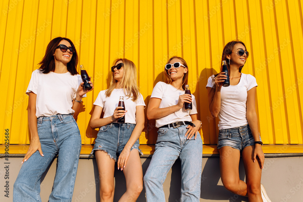 Obraz premium Smiling stylish young women in sunglasses with drinks standing against yellow wall in city on sunny summer day