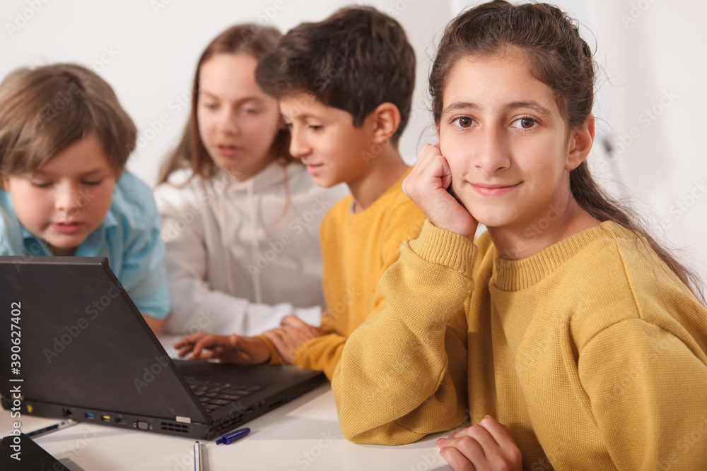 Cute Arab schoolgirl smiling to the camera, her friends using laptop at school on background Cute Arab schoolgirl smiling to the camera, her friends using laptop at school on background