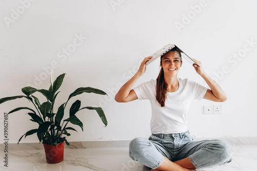 Happy woman smiling sitting on the floor making roof above her head with a laptop. Housewarming of single person. Moving to new flat alone. photo